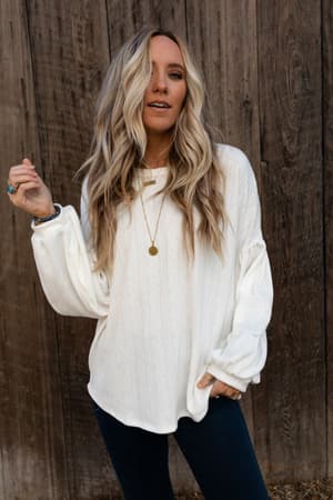 A woman with long, wavy blonde hair wears The Nest Sweet Heart Balloon Sleeve Knit Sweater in cream, dark jeans, and a gold necklace as she stands before a wooden wall, holding her hair and looking at the camera.