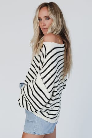 A woman with long blonde hair models the All My Stripes Oversized Sweater in cream and black with light denim shorts, standing against a plain white background and looking over her shoulder at the camera.