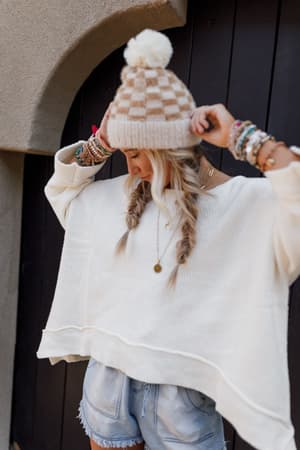 A woman with long blonde fishtail braids wears the Dana Oversized Diamond Texture Sweater in Ivory, denim shorts, a beige checkered knit beanie with a pom-pom, and colorful beaded bracelets near a dark wooden door.