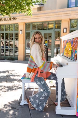 A woman with long blonde hair, wearing the Retro Babe Striped Cardigan - Cream and star-patterned jeans, smiles as she plays a colorful outdoor piano on a sunny sidewalk.