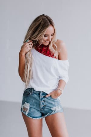 A woman with long blonde hair smiles while wearing a white off-shoulder top, ripped denim shorts, and the Baroque High Neck Velvet Bralette in Wine, standing against a plain light-colored background.