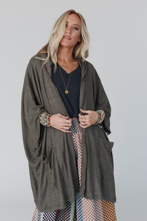 A woman with long blonde hair wears The Nest Boho Essential Hooded Ribbed Sweater in olive, styled oversized over a black top and colorful patterned skirt, accessorized with layered necklaces and bracelets against a light background.