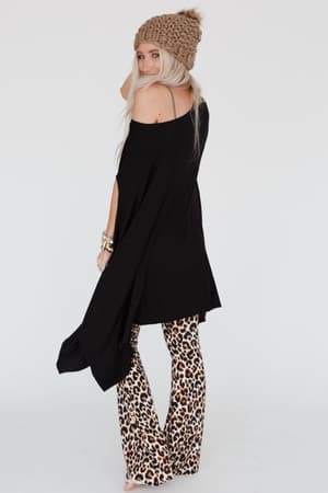 A woman with long blonde hair smiles over her shoulder against a plain white background, wearing The Wren Tunic - Black with a chunky brown knit beanie and leopard-print flared pants.