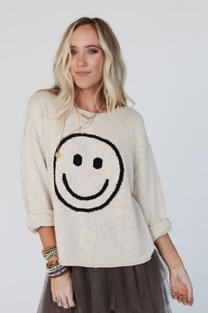 A woman with long, wavy blonde hair wears the Made You Smile Sweater in ivory, pairing the relaxed knit with stacked bracelets and a dark tulle skirt against a plain white background.