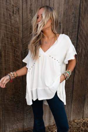 A woman with long blonde hair wears the More Than Meets The Eye Tunic Top in ivory and dark jeans, standing before a wooden wall, accessorized with multiple colorful bracelets on both wrists.