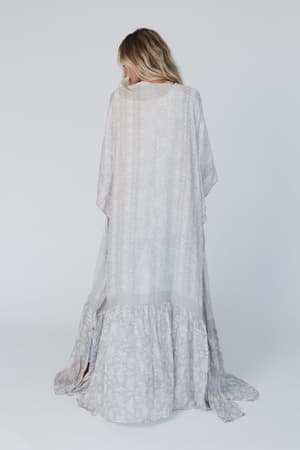 A person with long, wavy blonde hair faces away against a white background, wearing the Paisley Tapestry Free Flow Duster Kimono in gray with a ruffled hem.