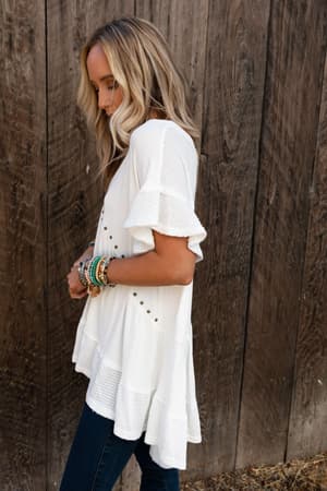 A woman with long blonde hair stands sideways against a wooden wall, looking down with her hands by her waist, wearing blue jeans and the More Than Meets The Eye Tunic Top in Ivory.