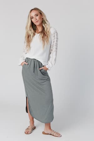 A woman with long blonde hair wears a white blouse and The Nest So Comfy Drawstring Maxi Skirt in Light Olive as she stands with her hands in her pockets against a plain white background.