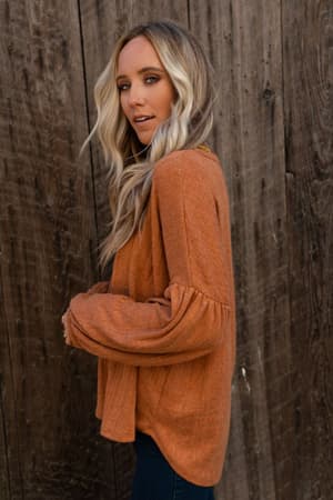 A woman with long blonde hair models The Nest Sweet Heart Balloon Sleeve Knit Sweater in burnt orange, standing sideways by a wooden wall and glancing over her shoulder at the camera.