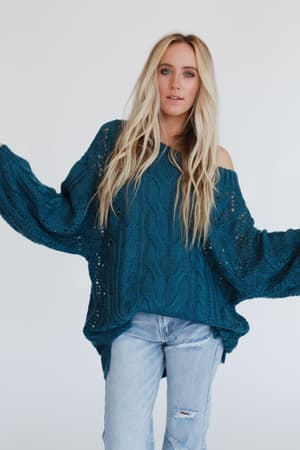 A person with long blonde hair wears the Golden Hour Bubble Sleeve Sweater Dress in teal, standing with arms raised and a neutral expression against a plain white background.