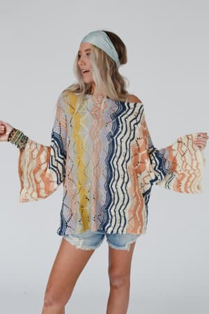 A woman in a light blue headband and the Rainbow Shores Knit Sweater - Multi with wavy patterns and distressed denim shorts stands against a plain background, holding her arms out to the sides.