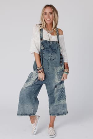 A woman wearing High Hopes Slouchy Denim Overalls - Denim Bandana, a loose off-shoulder white top, bracelets, and checkered slip-on shoes stands against a plain background, smiling.