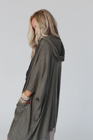 A woman with long blonde hair stands sideways, wearing The Nest Boho Essential Hooded Ribbed Sweater in olive. She pairs it with colorful beaded bracelets and rests her hand in the cardigan pocket against a plain light background.