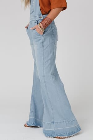 Wearing the Milly Distressed Hem Wide Leg Overall in denim with frayed hems, an orange ribbed top, and boho bracelets, the person has their hands tucked into the overall pockets; only their lower half is shown.
