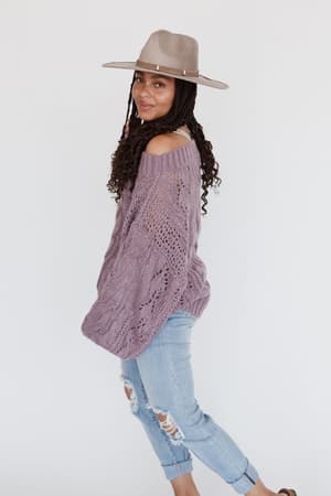 A woman with long braids smiles at the camera, wearing the Golden Hour Bubble Sleeve Sweater Dress in lavender, paired with a wide-brimmed hat and brown sandals, posing against a plain white background.