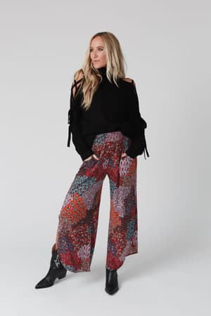 A woman with long blonde hair stands against a light gray background, hands in her pockets, wearing the Roxy Criss Cross Tie Shoulder Sweater in black, colorful wide-leg patterned pants, and black ankle boots.