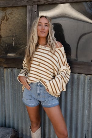 A woman with long blonde hair wears the All My Stripes Oversized Sweater in mustard, paired with denim shorts and white boots, standing confidently before a rustic corrugated metal and wooden wall.