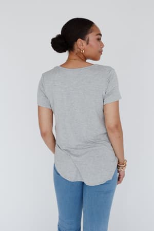A woman with dark hair in a bun, wearing the Scoop Neck Bralette Tee in gray and blue jeans, stands facing away from the camera against a white background. She accessorizes with gold hoop earrings and several bracelets.