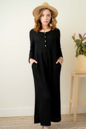 A woman in the Simply Comfort Jumpsuit - Black, featuring buttons and a scoop neck, stands indoors with hands in her pockets, wearing a tan hat. She poses by a light wall and wooden table with a potted plant, showcasing its relaxed silhouette.