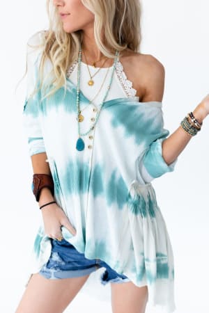Woman with long blonde hair poses in denim shorts, a blue and white tie-dye off-shoulder top, layered necklaces, bracelets, and the Light Of The Moon Crochet Trim Bralette in ivory against a white background.