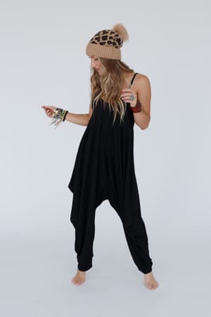 A woman smiles to the side, barefoot and accessorized with bracelets and a tan leopard-print beanie, wearing The Perfect Harem Jumpsuit - Black against a plain white background.