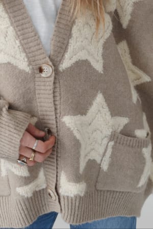 Wearing the Wrapped In Stars Oversized Cardigan in taupe, which features large white stars, a front pocket, ribbed cuffs, and button closure. This cozy piece is styled with eye-catching rings and blue jeans.