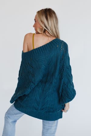 A woman with long blonde hair wears the Golden Hour Bubble Sleeve Sweater Dress in teal. She stands with her back to the camera, glancing to the side.