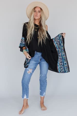 A woman in a wide-brimmed hat, barefoot and relaxed, wears the Fallon Embroidered Sleeve Poncho in black over distressed jeans, standing against a plain light background.