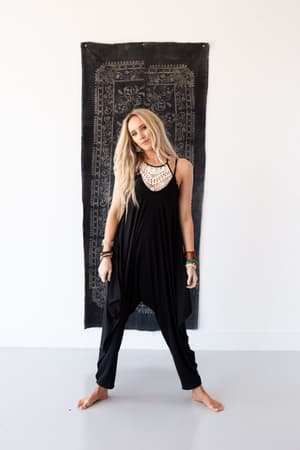 A woman with long blonde hair stands barefoot against a white wall and a decorative black textile, wearing The Perfect Harem Jumpsuit - Black, paired with layered necklaces and bracelets, striking a relaxed pose with a slight smile.