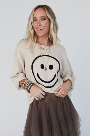 A woman with long blonde hair wears the Made You Smile Sweater - Ivory, layered necklaces, bracelets, rings, and a brown tulle skirt, smiling brightly against a plain white background.