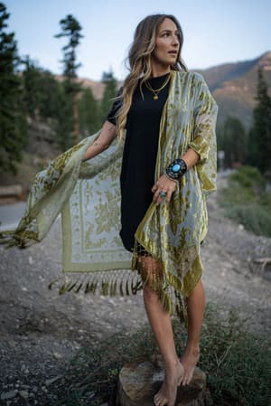 A barefoot woman stands outdoors on a tree stump, wearing the Crushin' On You Velvet Kimono - Olive over a black dress. She has long wavy hair, several bracelets, and is surrounded by mountains and trees.