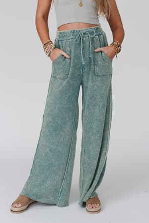 A person wears a light gray crop top with the Relaxing Robin Wide Leg Pant in New Teal, featuring front pockets and a drawstring waist, accessorized with multiple bracelets and tan platform sandals.