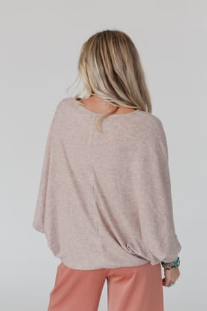 A woman with long blonde hair models The Nest Fly With Me Batwing Sleeve Knit Sweater in Sand, paired with peach pants, standing with her back to the camera against a plain light background.