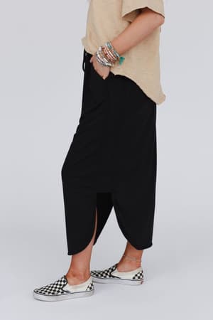 Person standing sideways wearing a loose beige t-shirt, The Nest So Comfy Drawstring Maxi Skirt - Black with side slits and elastic waistband, black-and-white checkered slip-on shoes, beaded bracelets on one arm, and hands in pockets.