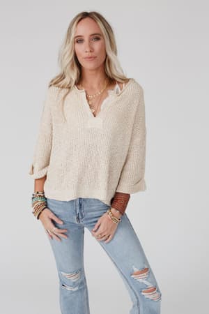 A woman with long blonde hair wears the Just Right Slouchy Sweater in oatmeal, layered necklaces, bracelets, and ripped light blue jeans as she stands against a plain light background, looking confidently at the camera.
