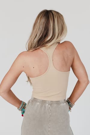 A woman with long blonde hair faces away from the camera, wearing a High Neck Racerback Brami in Nude and light gray pants, accessorized with multiple colorful beaded bracelets.