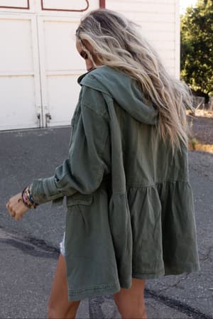 A person with long, wavy blonde hair wearing the Layers On Layers Perfect Boho Parka in olive walks outdoors on pavement near a white building, their back to the camera.