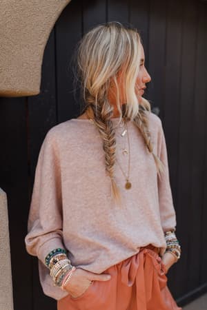 A woman with long blonde braids wears The Nest Fly With Me Batwing Sleeve Knit Sweater in Sand, rust-colored pants, layered necklaces, and colorful beaded bracelets, standing before a dark wooden door.