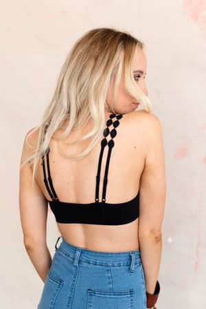 A woman with long blonde hair is shown from the back wearing the Imagine That Bralette - Black and high-waisted blue jeans, standing against a light textured background.