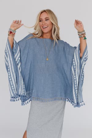 A smiling woman with long blonde hair wears the Fallon Embroidered Sleeve Tassel Poncho in blue, featuring patterned sleeves and tassels, paired with a light gray skirt and colorful bracelets against a plain background.
