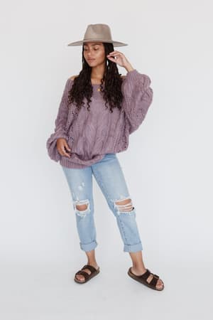 A woman with long curly hair poses against a white background, wearing the Golden Hour Bubble Sleeve Sweater Dress in Lavender, a wide-brimmed hat, ripped light blue jeans, and brown sandals. One hand lightly touches her hat.