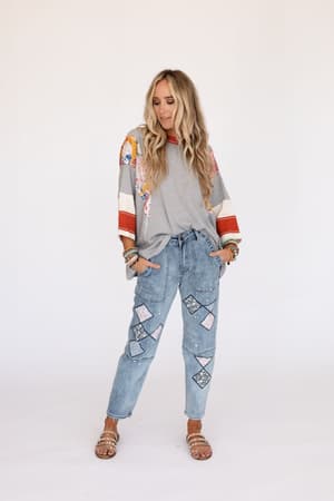 A woman with long blonde hair wears the Britt Patchwork Top in heather gray with colorful 3/4 sleeves, light blue jeans with geometric patches, sandals, and multiple bracelets, standing against a plain white background.