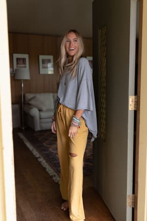 A woman with long blonde hair stands barefoot in a doorway, smiling. She wears The Nest Must Have High Low Tee in Heather Gray and mustard yellow ripped knee pants, accessorized with bracelets. A cozy living room is seen behind her.