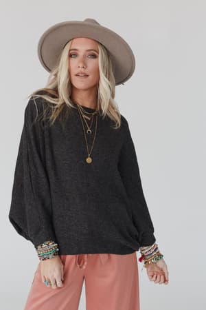 A woman with long blonde hair wears The Nest Fly With Me Batwing Sleeve Knit Sweater in charcoal, paired with a wide-brimmed hat, peach pants, layered necklaces, and colorful bracelets against a plain light background.