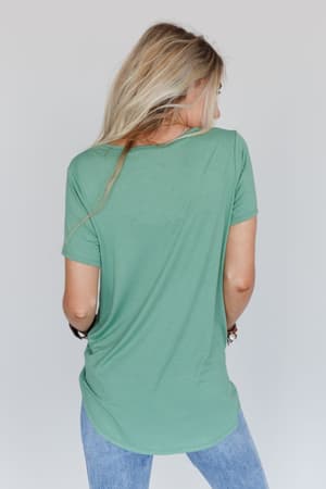 A person with long blonde hair stands with their back to the camera, wearing a loose Scoop Neck Bralette Tee in Sage and light blue jeans against a plain light background.