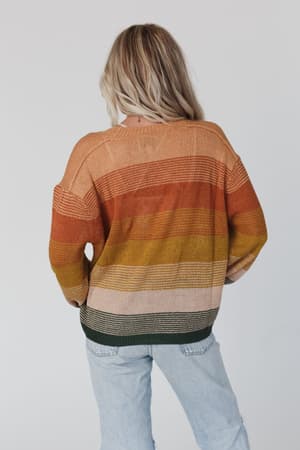 A person with long blonde hair is seen from behind wearing the Going Places Gradient Stripe Sweater - Mustard with orange, yellow, green, and beige stripes and light blue ripped jeans against a plain light background.