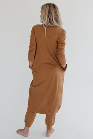 A barefoot person with long blonde hair stands facing away, wearing the Set to Go Textured Jumpsuit And Cardigan Set in camel, featuring a loose, long-sleeved cardigan over matching jumpsuit pants against a plain white background.