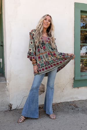 A woman with long blonde hair stands outside, smiling as she holds out one side of her Leona Floral Print Tunic - Sage, paired with flared blue jeans and studded sandals. A light wall and a green-framed window are behind her.
