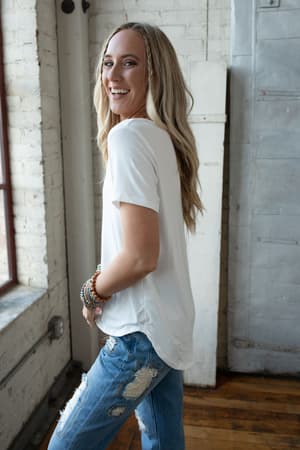 A woman with long blonde hair, wearing the Scoop Neck Bralette Tee in white and ripped blue jeans, stands by a window indoors, smiling over her shoulder. She accessorizes with beaded bracelets in a bright, industrial-style space.