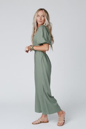 A woman with long blonde hair wears the Weekender Puff Sleeve Jumpsuit in olive, featuring wide legs, styled with flat studded sandals and layered bracelets. She stands against a plain light background.